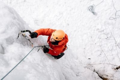 Person klettert mit Helm und Eispickel an gefrorenem Wasserfall; Seil zur Sicherung sichtbar.