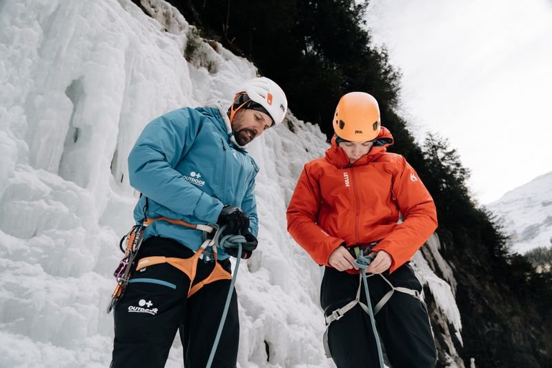 Zwei Personen mit Helmen und Klettergurten stehen vor einer vereisten Felswand in den Bergen.