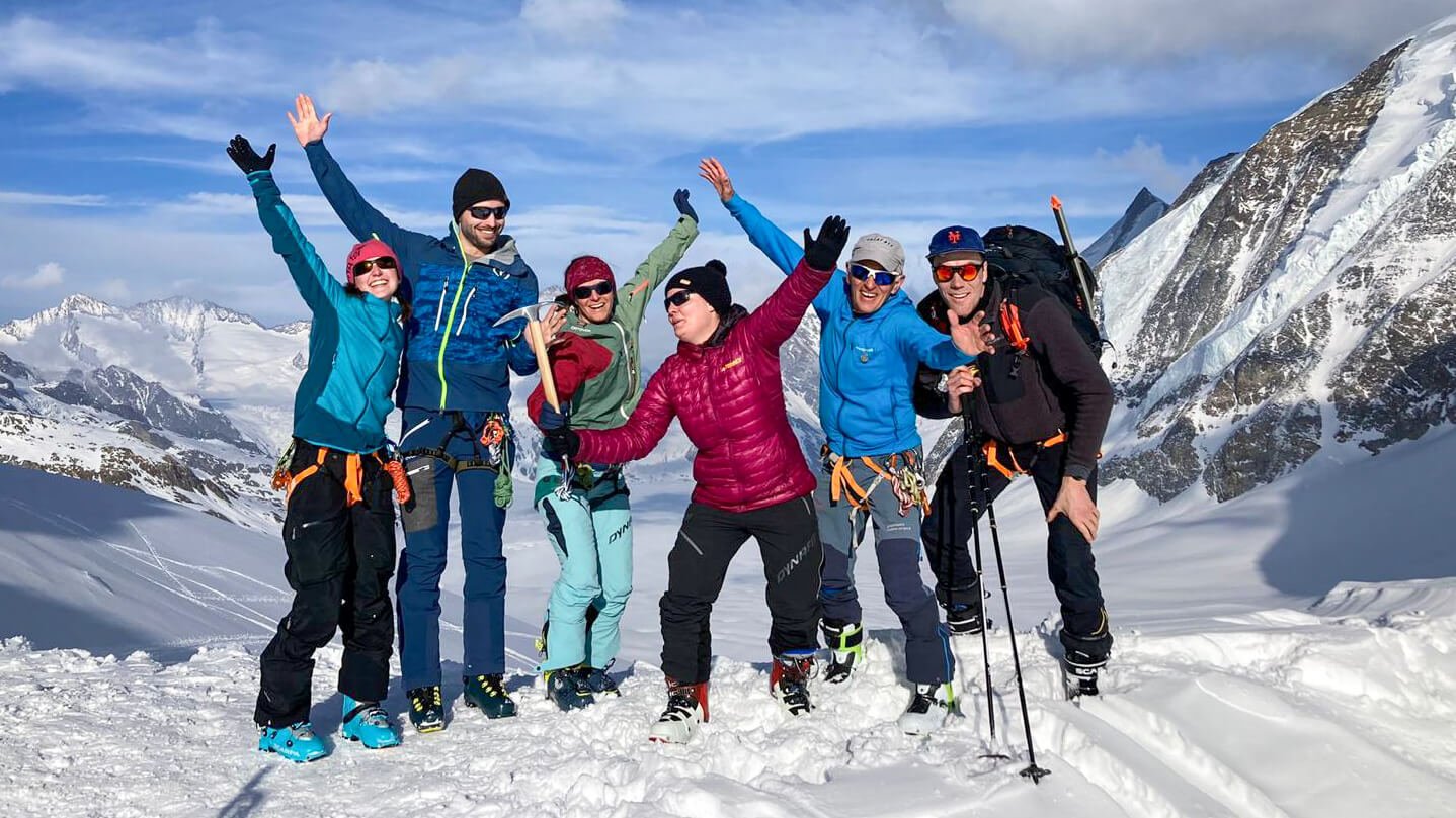 Gruppe von Personen mit Kletterausrüstung und Skistöcken auf schneebedecktem Berggipfel.