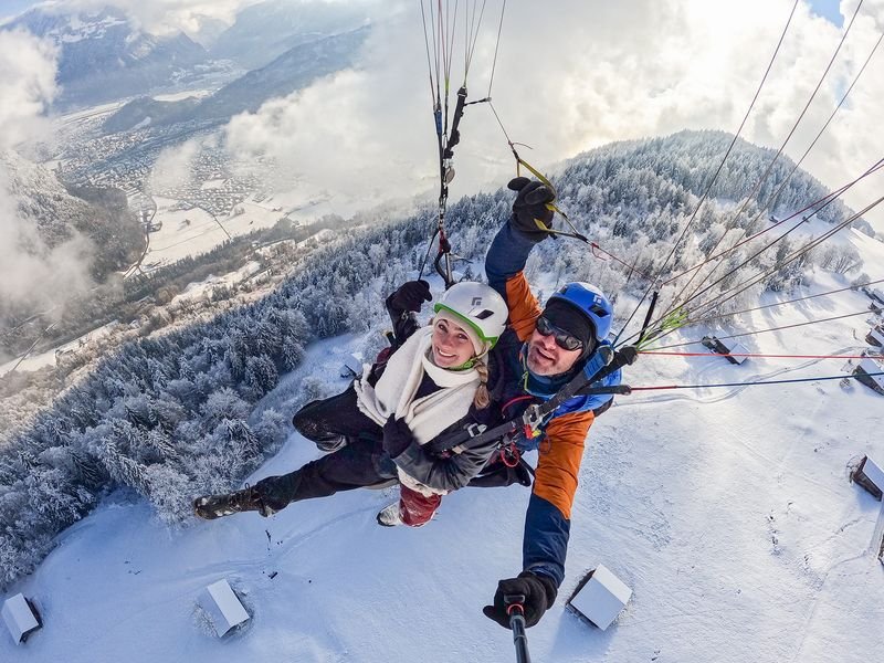 Two people tandem paragliding over snowy mountains and a valley.
