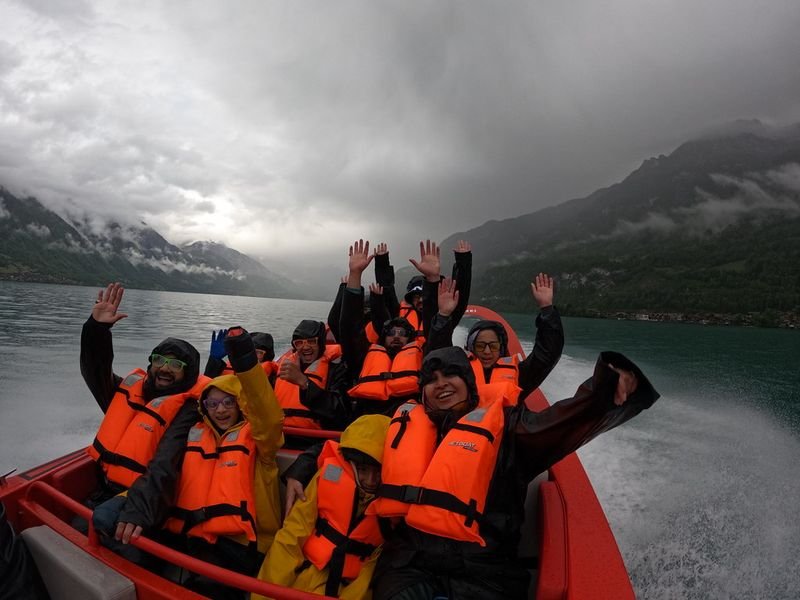 Gruppe von Personen mit orangefarbenen Schwimmwesten auf einem Schnellboot auf einem See, umgeben von Bergen und Wolken.