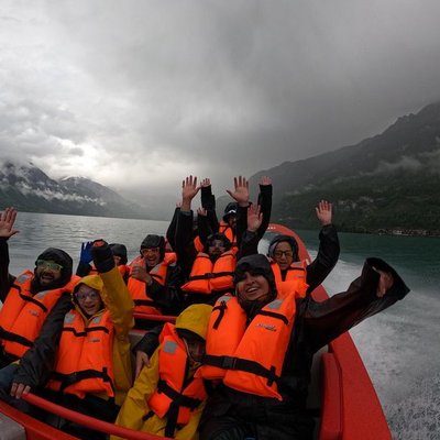 Gruppe von Personen mit orangefarbenen Schwimmwesten auf einem Schnellboot auf einem See, umgeben von Bergen und Wolken.