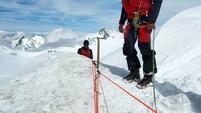 People with climbing equipment and ropes on a snow-covered mountain peak, surrounded by the Alps.