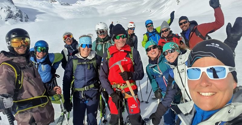 Group of people with ski equipment and helmets posing in the snowy mountain area.