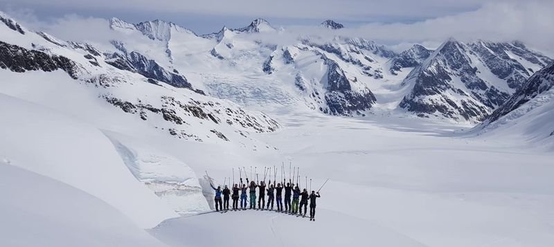 Group of skiers with poles on a snow-covered mountain peak, surrounded by an Alpine panorama.
