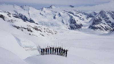 Group of skiers with poles on a snow-covered mountain peak, surrounded by an Alpine panorama.