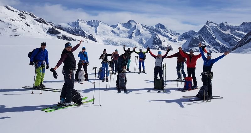 Group of skiers with helmets and poles on a snow-covered mountain with an Alpine backdrop.