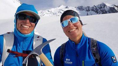Two people in blue outdoor clothing with sunglasses and ice axes in snow-covered mountains.