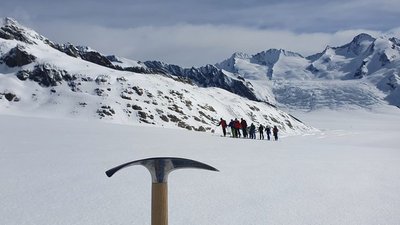 Group of people with mountaineering equipment on a snowy glacier, ice axe in the foreground.
