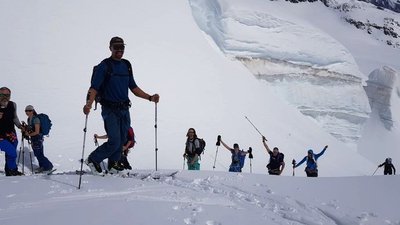 Group of people with sticks hiking in the snow in front of a glacier wall.