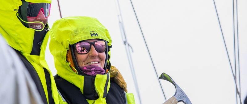 People in yellow jackets and sunglasses on a sailboat, holding a sail tool.