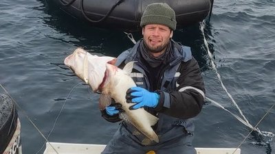 Person in outdoor clothing holding a large fish on a boat, with water and an inflatable boat in the background.