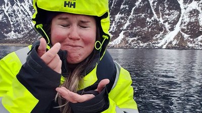 A person in a yellow jacket on a boat in front of snow-covered mountains and water.