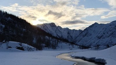 Snow-covered mountain landscape with a river, surrounded by high mountains and cloudy sky at sunset.
