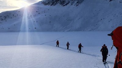 People in red jackets are hiking with ski poles through a snowy mountain landscape at sunrise.
