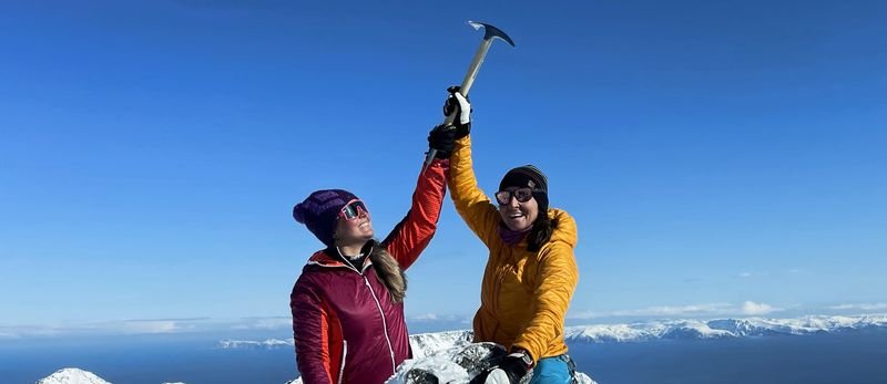 Two people in winter clothing on a snowy mountain peak, holding ice axes in the air.