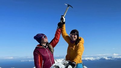 Two people in winter clothing on a snowy mountain peak, holding ice axes in the air.
