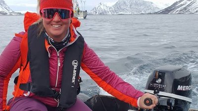 Person in boat with life jacket and cap is steering motorboat on the lake, surrounded by snow-covered mountains.