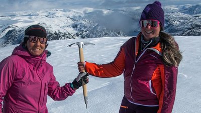 Two people in winter clothing with sunglasses and ice axes on a snow-covered mountain peak.