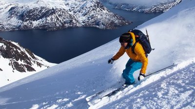 Person skiing down a snow-covered slope, with backpack and helmet, mountain landscape in the background.