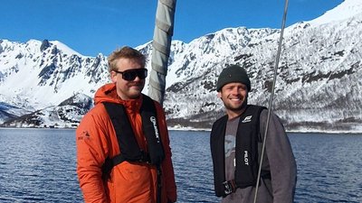 Two people with life jackets on a boat in front of snow-covered mountains and a lake.