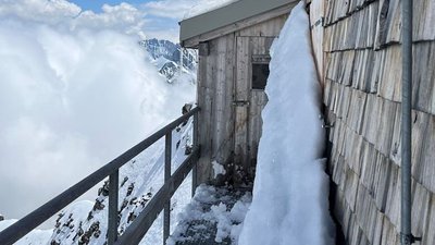 Wooden cabin in snowy mountains with snowdrifts, clouds, and mountain peaks in the background.
