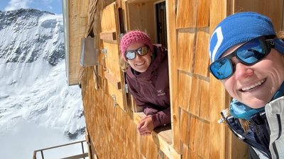 Two people with sunglasses are looking out of a wooden window at the snow-covered mountains.