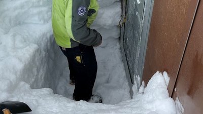 Person in a green jacket stands in the snow in front of a metal door, surrounded by high snow walls.