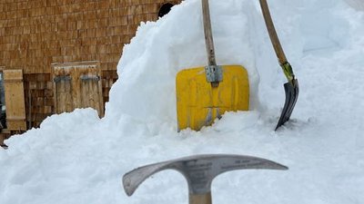 Ice pick and shovel are stuck in the snow in front of a mountain hut with a wooden facade.
