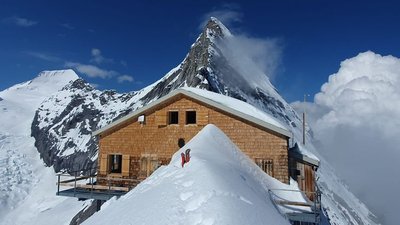Wooden cabin on a snow-covered mountain peak with surrounding snowy mountains in the background.