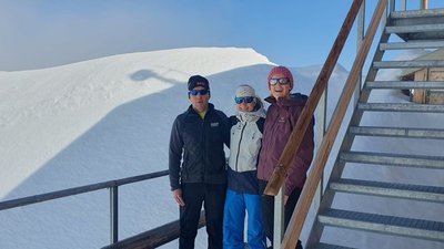 Three people in winter clothing with sunglasses are standing on a wooden platform in the snow.