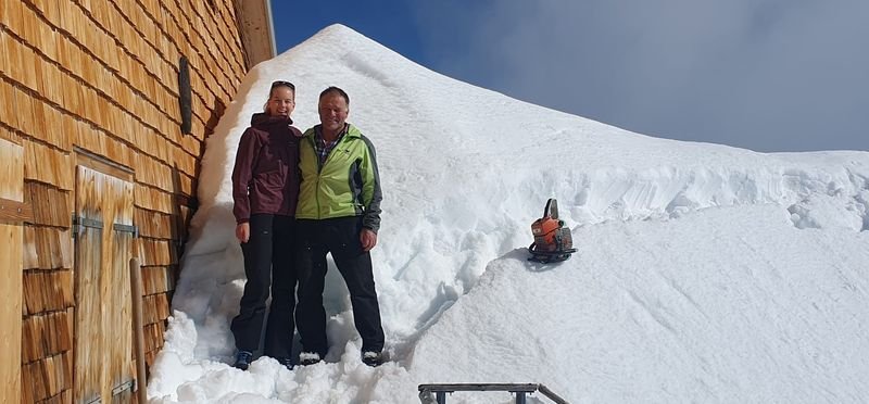 Two people are standing in winter clothing next to a snow-covered building in the mountains.