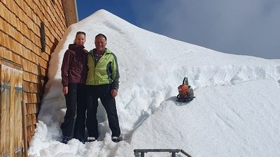Two people are standing in winter clothing next to a snow-covered building in the mountains.