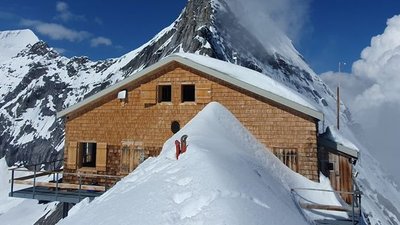 Wooden cabin in snowy mountains, person in red jacket standing on snow, blue sky in the background.