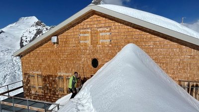Person in front of a mountain hut in the snow, surrounded by snow-covered mountains, in sunny weather.