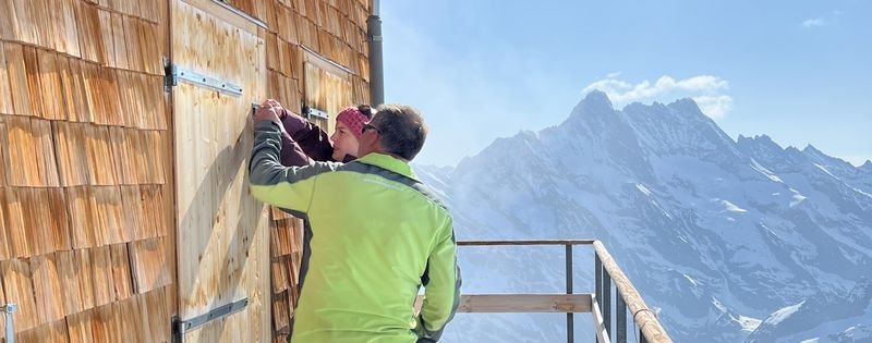 People in outdoor clothing are opening a wooden door at a mountain hut, with snow-covered mountains in the background.