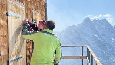 People in outdoor clothing are opening a wooden door at a mountain hut, with snow-covered mountains in the background.