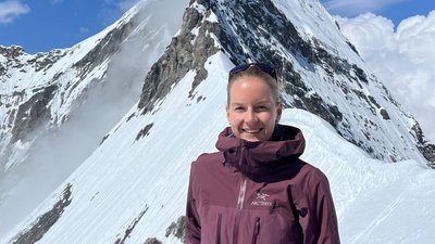 Person in purple jacket stands in front of snow-covered mountain in the Alps, blue sky in the background.