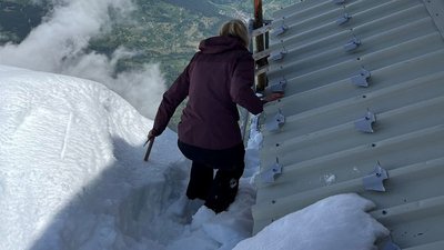 Person on snow-covered mountain next to cabin, holding onto railing.