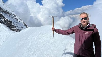 Person on snow-covered mountain with sunglasses and ice axe, surrounded by clouds.