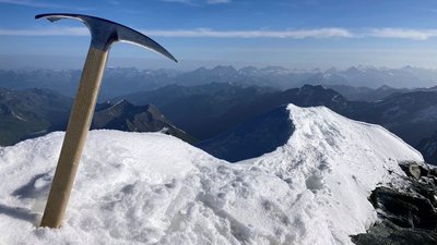 An ice axe is stuck in the snow on a mountain peak overlooking a mountain landscape.