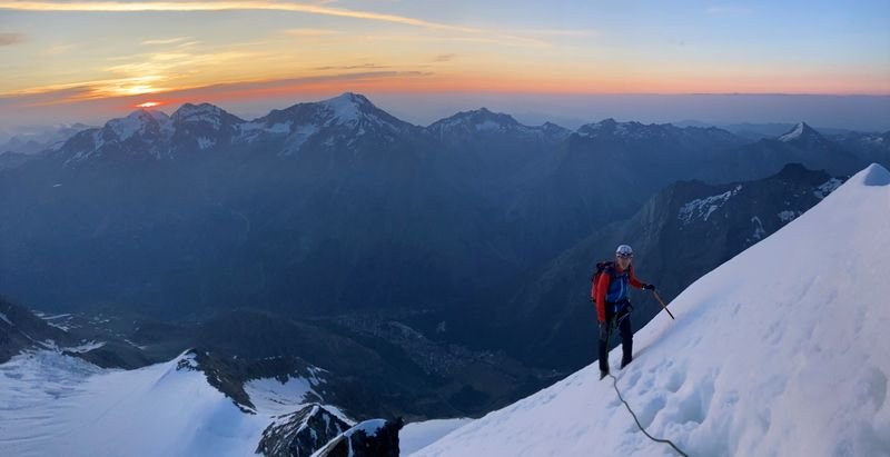 Mountaineer with helmet and rope on a snow-covered slope, with mountains in the background at sunrise.