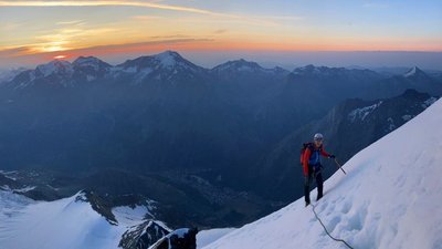 Mountaineer with helmet and rope on a snow-covered slope, with mountains in the background at sunrise.