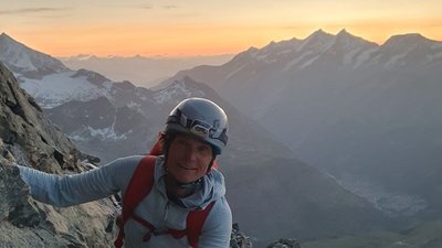 Person with helmet and climbing equipment on a rock wall, with mountains in the background during sunset.