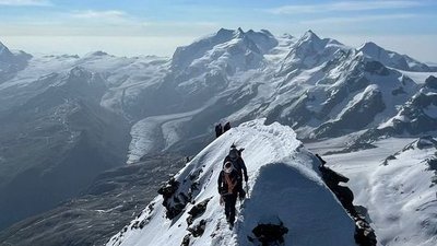 Mountaineers with helmets and ropes on a snow-covered ridge in the Alps, surrounded by mountains.