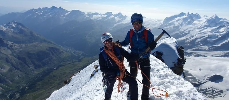 Two people with helmets and climbing gear are standing on a snow-covered mountain peak in the Alps.