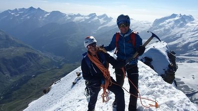 Two people with helmets and climbing gear are standing on a snow-covered mountain peak in the Alps.
