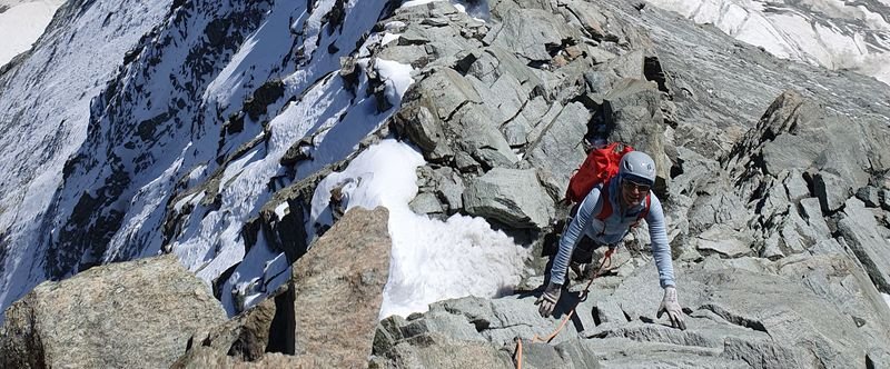 Climber with helmet and backpack on rocky mountain ridge, snow-covered mountains in the background.