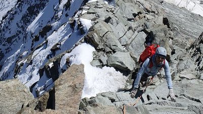 Climber with helmet and backpack on rocky mountain ridge, snow-covered mountains in the background.