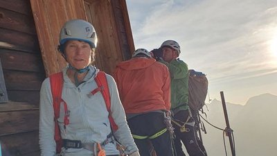 People with helmets and climbing harnesses in front of a wooden hut in a mountain landscape at sunrise.